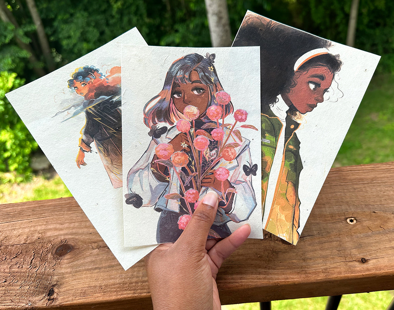 Hand holding three GDBee art prints featuring diverse characters outdoors on wooden railing